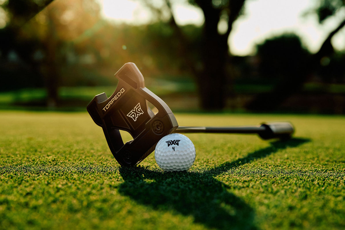 A Torqueed golf putter head resting on a golf green next to a white golf ball, with a blurred outdoor golf course background and warm sunlight.